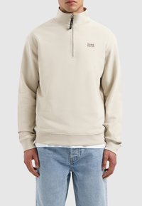 Beige ritsvest met hoge kraag, geribde manchetten en zoom, met het "PURE PATH" logo in bruin op de linkerkant van de borst. Zachte textuur.