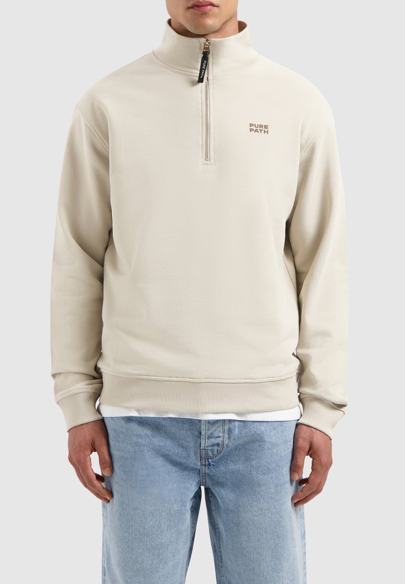 Beige ritsvest met hoge kraag, geribde manchetten en zoom, met het "PURE PATH" logo in bruin op de linkerkant van de borst. Zachte textuur.