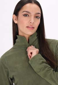 Pullover in pile verde oliva con colletto alto e chiusura a mezza zip. Tessuto morbido, maniche lunghe e dettagli cuciti sulle cuciture.