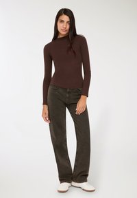 Kobieta z długimi ciemnymi włosami, ubrana w dopasowany brązowy top z długim rękawem, luźne ciemne dżinsy i białe trampki, stojąca na jednolitym jasnym tle.