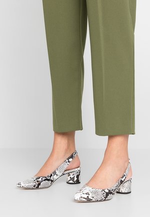 Jalat, joissa valkoiset ja mustat käärmeenahkaiset slingback-pystykorkokengät, yhdistetty oliivinvihreisiin lyhytlahkeisiin housuihin.
