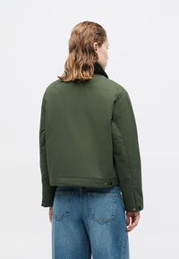 Groene bomberjack met een zwarte kraag, gemaakt van een gladde stof. Heeft een cropped ontwerp en knoopsluitingen aan de onderkant. Gecombineerd met blauwe jeans.