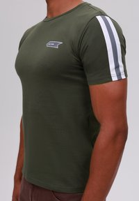 Camiseta ajustada de color verde oliva hecha de una tela suave, con dos rayas blancas y grises en las mangas cortas y un pequeño logo en el pecho.