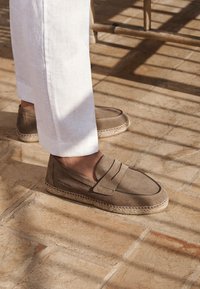 Bruine suède instapschoenen met gestikte accenten en jute details, gecombineerd met witte linnen broeken, op een gestructureerde vloer.