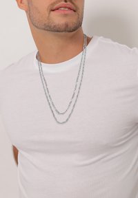 Sølvfarvede, lagdelte kædedesigns med varierende linkmønstre, båret over en simpel hvid t-shirt. Fokuser på den glatte metalliske finish og struktur.