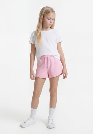 Jeune fille debout, portant une chemise blanche à manches courtes, un short rose avec cordon, des chaussettes blanches et des baskets blanches sur un fond blanc.