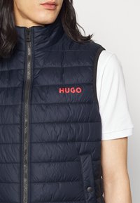 Chaleco acolchado azul marino con un alto cuello, con secciones acolchadas en horizontal y un logotipo rojo "HUGO" en el pecho. Cierre frontal con cremallera.