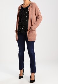 Cardigan lavorato a maglia di un rosa chiaro con apertura frontale e due tasche, indossato sopra una maglietta nera con pois bianchi, abbinato a jeans blu scuro e tacchi neri.