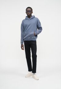 Moleton azul com um bolso frontal, combinado com calças pretas slim-fit e sapatos beje sem atacadores. O modelo está de pé contra um fundo branco.