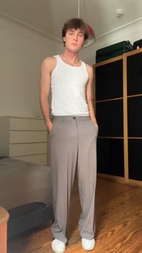 Lys grå, plisserte bukser med rett ben-design, båret med en hvit tanktop og hvite joggesko, med hendene i lommene, nøytral bakgrunn.