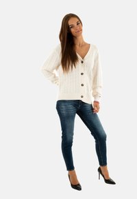 Giovane donna con capelli lunghi, che indossa un cardigan bianco con bottoni, jeans skinny blu e scarpe con tacco alto nere, in posa con una mano sulla vita.