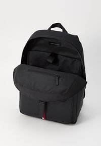 Sac à dos en tissu noir avec un rabat supérieur et une poche zippée à l'avant. L'intérieur comprend un compartiment principal et une étiquette de marque sur la doublure.