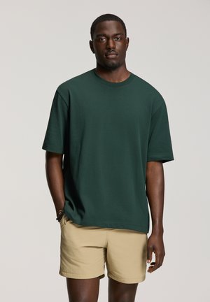 Man draagt een donkergroen oversized T-shirt en beige korte broek, staat met één hand in zijn zak tegen een effen achtergrond.