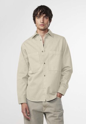 Light beige button-up shirt made of cotton, featuring two chest pockets, a collar, and long sleeves. Paired with matching beige pants.