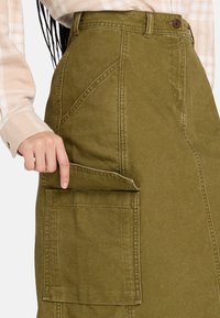 Falda de algodón verde oliva hasta la rodilla, con un bolsillo frontal y costuras estructuradas. La textura es suave con un ligero brillo.
