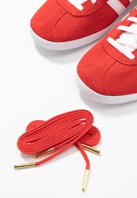 Röda mockaskor med vit gummisula och snören. Inkluderar extra röda snören med guldfästen, rullade bredvid skorna.