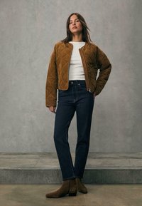 Jaket od debelog kordon materijala smeđe boje sa prednjim zipom, bijela košulja, tamnoplave traperice i smeđi čizmi do gležnja s niskom petom.