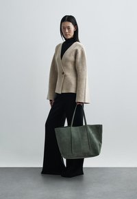 Beige jas van wolmix met wijde mouwen, gecombineerd met een zwarte broek. Model draagt een grote groene leren totebag met twee lange hengsels.