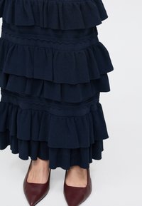 Φούστα σε ναυτικό μπλε με στρώσεις βολάν συνοδευόμενη από μπορντό γόβες με μυτερή μύτη σε λευκό φόντο.