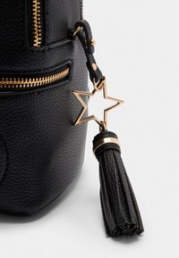 Schwarze strukturierte Tasche mit goldenen Beschlägen, ausgestattet mit einem sternförmigen Anhänger und einem schwarzen Quasten-Detail.