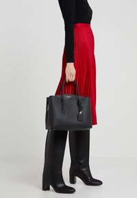 Borsa in pelle nera a grana con doppio manico, chiusura con cerniera, dettagli in metallo dorato e un'etichetta per bagagli, mostrata con una gonna rossa plissettata.