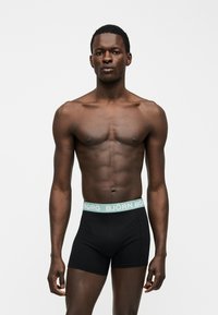 Άνδρας μοντέλο με σκούρο δέρμα που φοράει μαύρα μποξεράκια με ανοιχτό μπλε μέση όπου αναγράφεται «BJÖRN BORG», στέκεται μπροστά σε απλό λευκό φόντο.