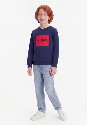 Enfant aux cheveux roux portant un sweat-shirt bleu marine avec le logo rouge "HUGO", un jean bleu clair et des baskets blanches, debout et souriant.