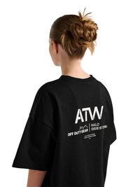 Kobieta o brązowych włosach związanych w kok, ubrana w luźną czarną koszulkę z białym napisem i grafiką na plecach na białym tle.