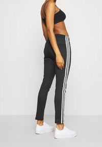 Mujer vistiendo pantalones deportivos negros con franjas laterales blancas, sujetador deportivo negro y zapatillas blancas, de pie contra un fondo claro y liso.