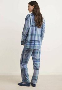 Sininen ruudullinen pyjamapukusetti, joka on valmistettu pehmeästä kankaasta. Se sisältää löysän napillisen paidan ja siihen sopivat housut, joissa on monia sinisen sävyjä ja kuvioita.
