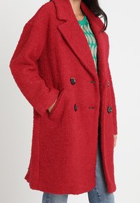 Rød, oversized dobbeltradet frakke lavet af tekstureret stof, med store sorte knapper og sidelommer, båret over en grøn top.