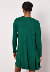 Abito a maglia verde a coste con maniche lunghe, due tasche laterali e orlo svasato. Il tessuto sembra morbido e testurizzato.