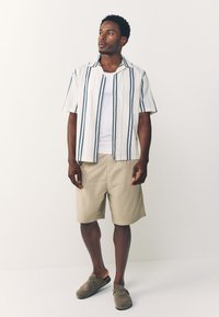 Chemise blanche à rayures bleues, manches courtes, short beige et chaussures grises à enfiler. Design simple, matière en coton, coupe décontractée.