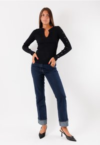 Top nero a maniche lunghe con scollatura a V e dettagli di bottoni lungo le maniche, abbinato a jeans in denim scuro risvoltati alle caviglie e décolleté neri a punta.