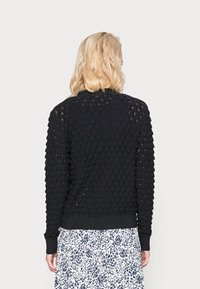 Woman with blonde hair wears a black textured long-sleeve sweater and a white skirt with black abstract spots, shown from the back.