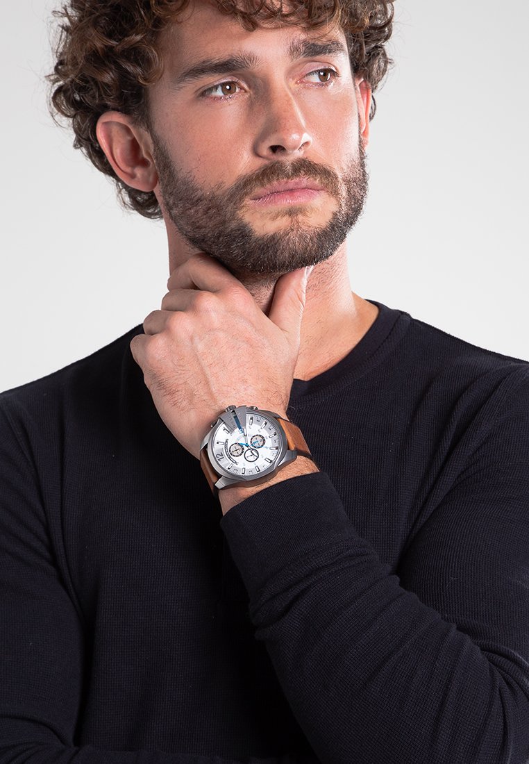 Montre argentée avec cadran blanc, accents bleus et bracelet en cuir marron. Portée au poignet d'un homme, la main placée de manière réfléchie contre le menton.