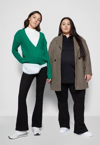 Duas pessoas estão lado a lado. Uma veste um cardigan verde de malha em forma de cables por cima de uma camisola de gola alta branca, e a outra usa uma blusa preta e um casaco de padrão pied-de-poule. Ambas têm calças pretas ajustadas e ténis brancos.