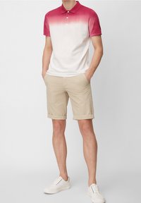 Polo shirt pour hommes en blanc, dégradé vers le rouge au col, avec des manches courtes. Associé à un short beige et des baskets blanches.
