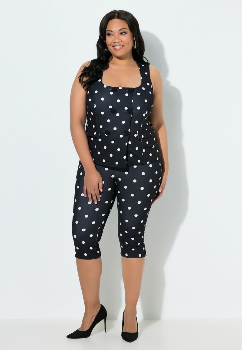 Femme souriante aux longs cheveux noirs porte un gilet sans manches à pois noir assorti à un pantalon court et des talons hauts noirs, debout contre un mur blanc.