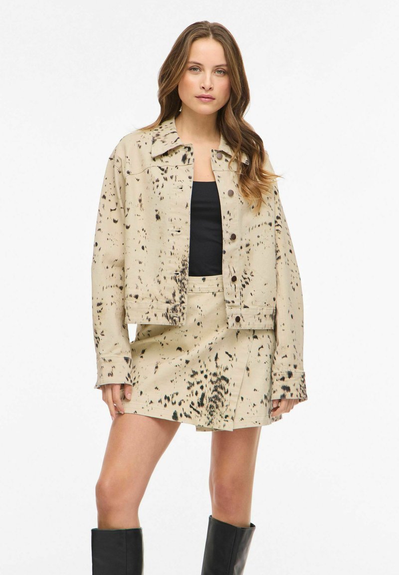 Bež teksas jakna s crnim mrljama, s ovratnikom i prednjim zatvaranjem na kopče, u kombinaciji s odgovarajućom suknjom.