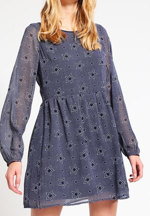 Femme portant une robe longue à manches longues bleu marine transparent avec des motifs d'étoiles blanches et une taille froncée.