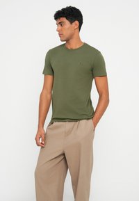 T-shirt din bumbac verde măsliniu, cu guler rotund, mâneci scurte și un mic logo brodat pe piept. Asortat cu pantaloni bej, largi.