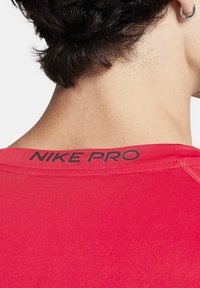 Rød sportsskjorte med korte ærmer, lav udskæring og sort "NIKE PRO" logo på ryggen. Glat, strækbart stof med synlige sømme.