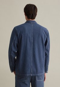 Blue striped denim jacket with a collar, long sleeves, button cuffs, and back seam detailing. The fabric has a textured finish.