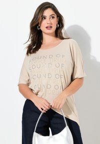 Beige T-shirt med en løs pasform og bred hals, prydet med et metallic tekstmønster. Sammen med mørkeblå bukser og en sølvfarvet håndtaske.