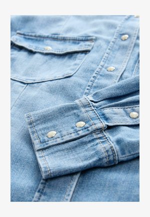 Camisa de mezclilla azul claro con botones de presión plateados, dos bolsillos en el pecho y costuras en contraste. La tela presenta una superficie suave y texturizada.