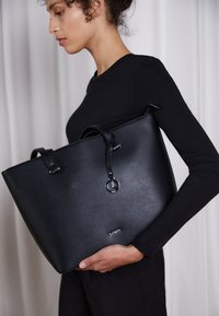 Schwarze Leder-Tote-Bag mit strukturierten Oberflächen, zwei oberen Griffen, kreisförmigem Hardware-Detail und Logo auf der Vorderseite. Rechteckige Form, geräumiges Design.