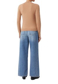 Frau in einem sandfarbigen Pullover und hellblauen, weiten Jeans, stehend mit dem Rücken zur Kamera.