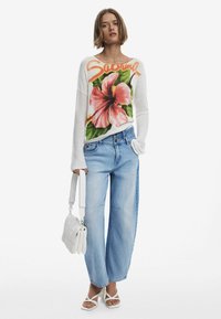 Maglione bianco a maglia con un design di ibisco rosa e il testo "Sabrina", abbinato a pantaloni in jeans azzurri a gamba larga e sandali bianchi con cinturini. Tiene una borsa bianca.
