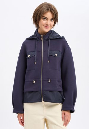 Jeune femme portant une veste bleu marine zippée avec capuche à cordon et un pantalon beige, souriante sur fond blanc uni.